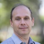 Louis is wearing a blue shirt, standing outside and smiling at the camera