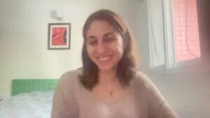 Natasha has dark shoulder length hair and is wearing a neutral colored top while sitting in her bedroom and smiling at the camera.