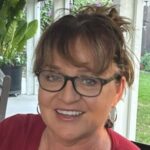 Susan has short brunette hair and is wearing glasses and a red top as she smiles at the camera