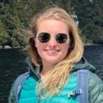 Clare has long blonde hair and is wearing sunglasses while standing outside with a body of water behind her