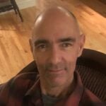 Dion is a man sitting in a chair and smiling at the camera. He is wearing a dark collared shirt.