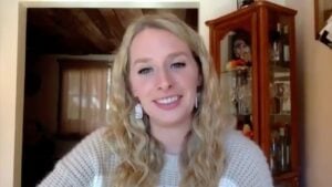 Courtney has long, fair, wavy hair. She is smiling at the camera while sitting inside her house.