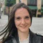 Sabina is in an outdoors urban environment and smiling at the camera. She is wearing a dark jacket and has long dark hair.