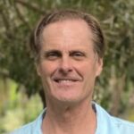 Portrait of a middle-aged man outdoors, smiling gently at the camera with trees softly blurred in the background