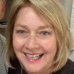 Close-up portrait of a smiling woman with blonde hair, looking directly at the camera indoors.