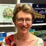 Portrait of a smiling woman wearing glasses, standing indoors in front of a bookshelf with colorful books in the background.