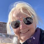 Smiling woman wearing colorful sunglasses outdoors on a sunny day, holding a drink with a clear blue sky and building in the background.