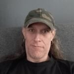 Person wearing a camouflage baseball cap and black shirt, looking at the camera while seated against a gray background.