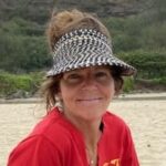 Smiling woman wearing a patterned sun visor and red shirt outdoors near a beach.