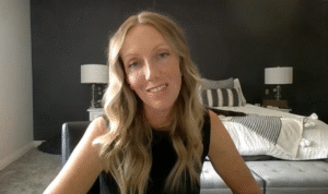 Woman smiling during a video interview in a bedroom with a neatly made bed and lamps in the background.