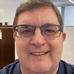 Close-up photo of a smiling man wearing rectangular glasses in an office setting.