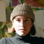 Close-up portrait of a woman wearing a gray knit hat and black turtleneck, with sunlight across her face.