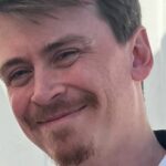 Close-up portrait of a smiling man with short brown hair, a mustache, and a goatee.